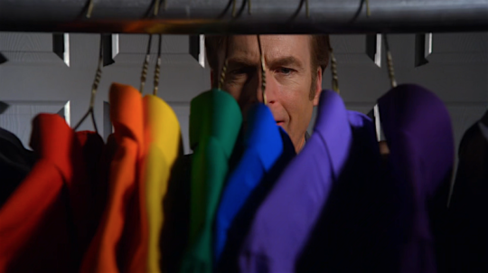 A picture of an excited man's (fictional character Jimmy McGill) staring at a set of suits, each of which spurs a unique colour (red, orange, yellow, green, blue, and purple in that order from left to right)