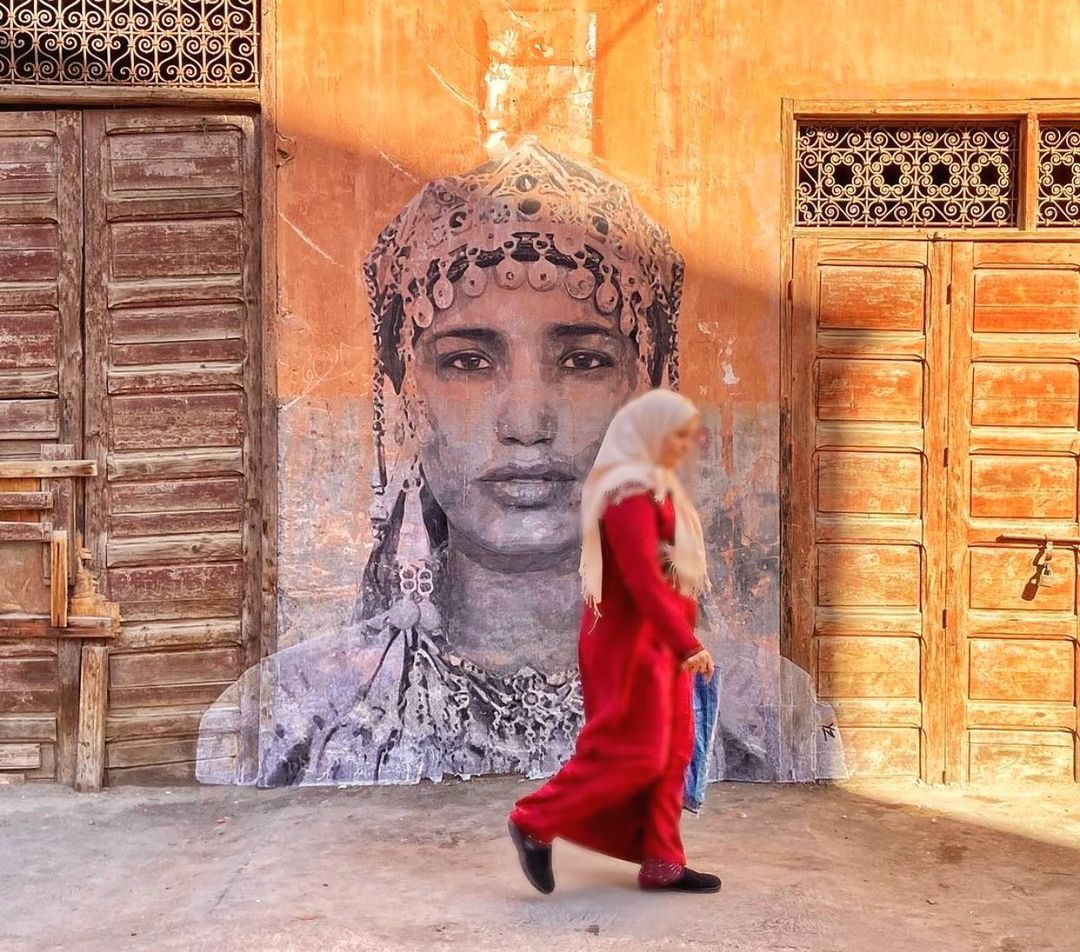 A woman walking in front of a wall with art of an Amazigh woman on it.