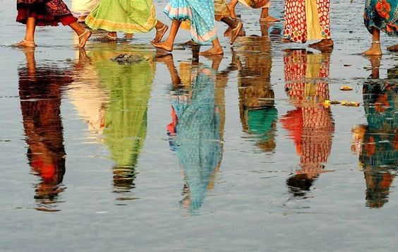 A group of women in their cultural clothing walking along the beach with their reflections shown through the water